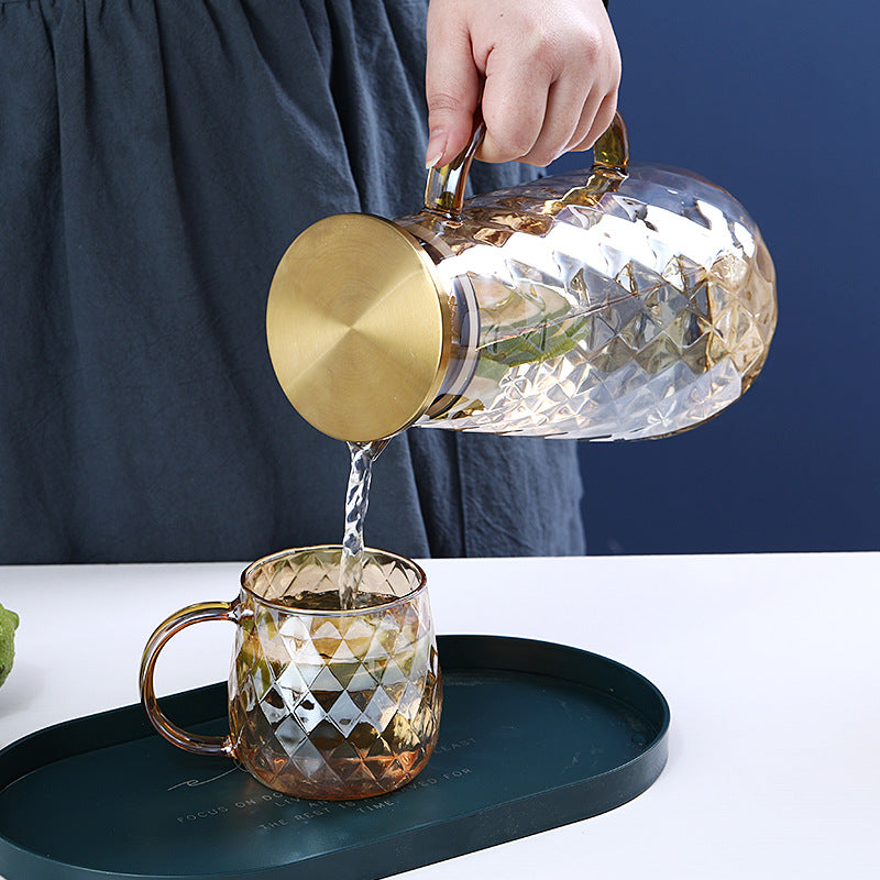 Glass teapot with gold accents being poured into a matching glass mug on a white surface.