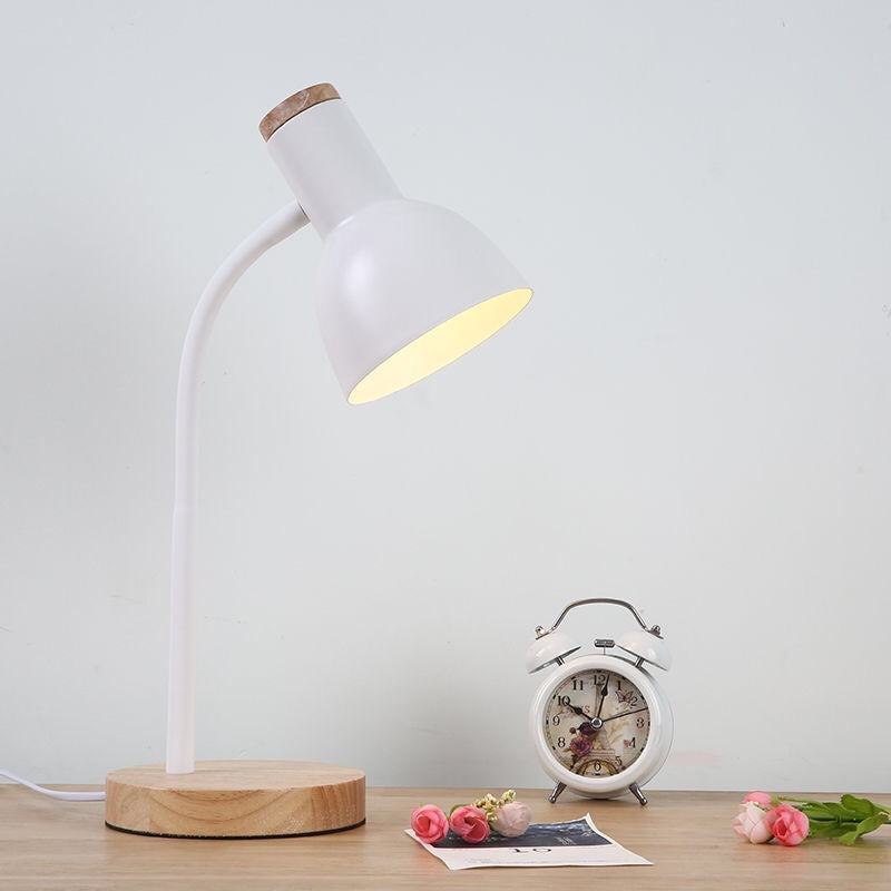 White desk lamp with wooden base on a table with a white alarm clock and flowers.