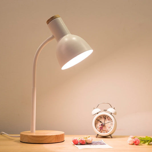 White desk lamp with wooden base on a table next to a small clock and flowers.